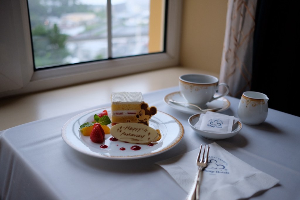 窓辺に並べられた記念日のケーキと紅茶のセット。皿にはカラフルなフルーツと「Happy Anniversary」と書かれたデザートが添えられている。