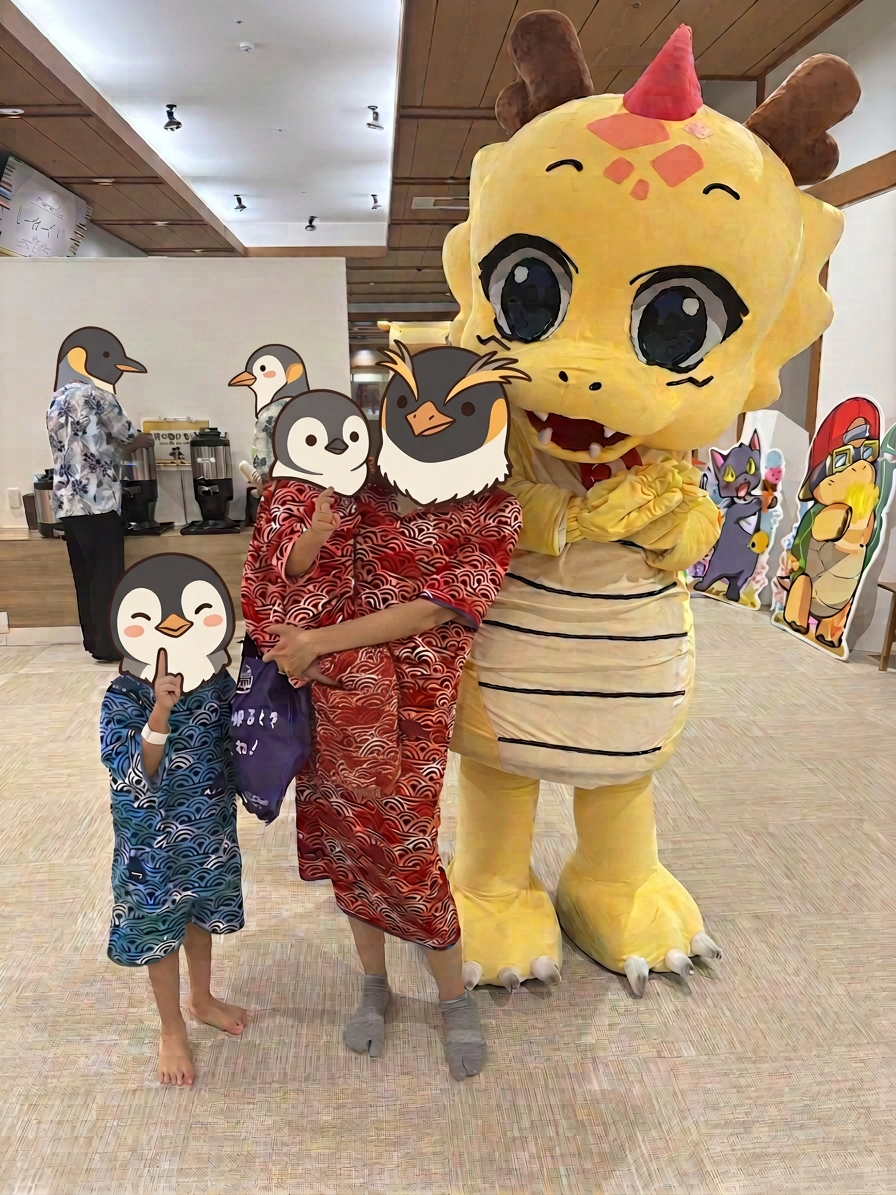 三日月キャラクター カラフルな浴衣を着た子供たちと大きなドラゴンの着ぐるみのキャラクターが一緒にポーズをとっている写真。背景には他のキャラクターの立て看板が見える。