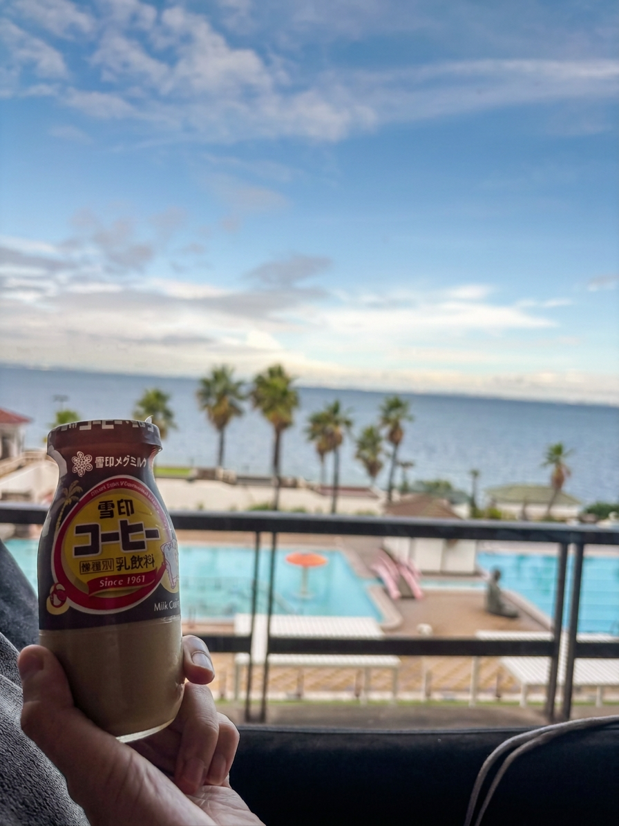 img_4351 窓の外に海とプールが見える景色を背景に、コーヒー飲料の瓶を持つ手