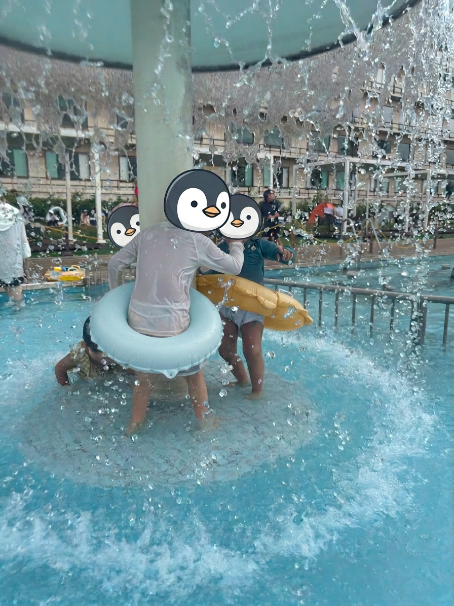 img_4226 子供たちが水の噴水の下で遊んでいる様子。顔にはペンギンのデザインのマスクを着用している。