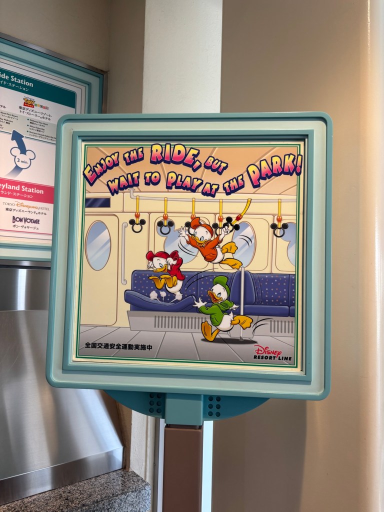 ディズニーリゾートラインのサイン。キャラクターたちが電車の中で楽しんでいる様子を描いており、「乗って楽しんで、でも公園で遊ぶのを待ってね！」と書かれている。