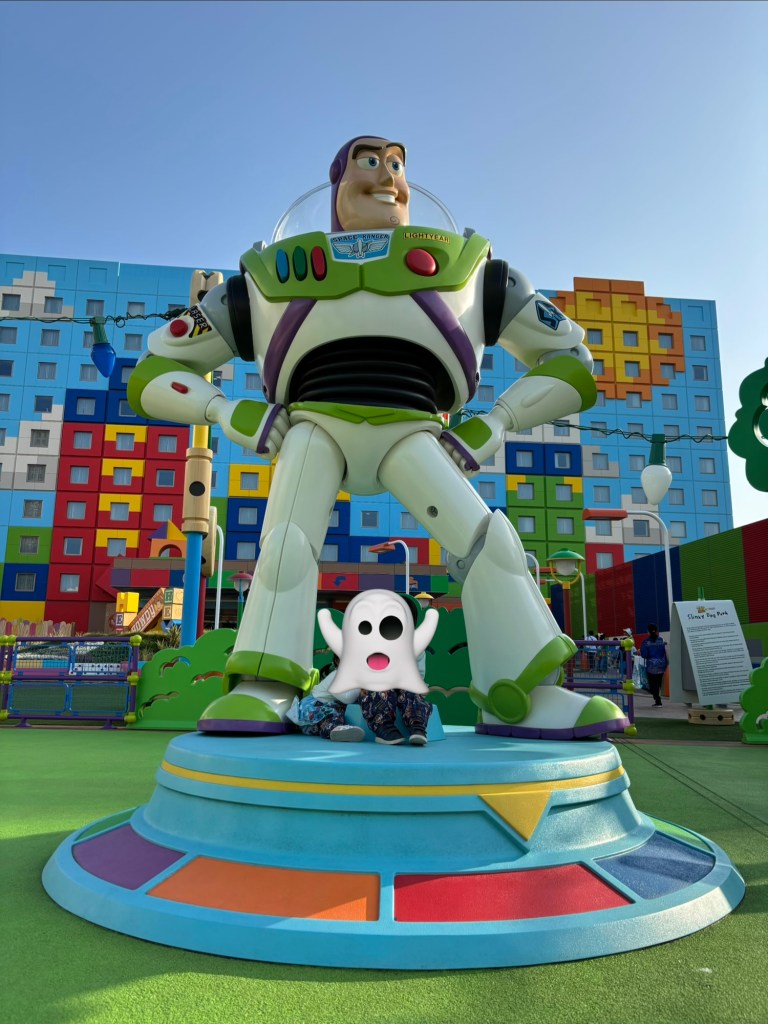 バズ・ライトイヤーの巨大な立像の前に座っている子供とゴーストのマスコット。背景にはカラフルな建物が見える屋外の遊び場。