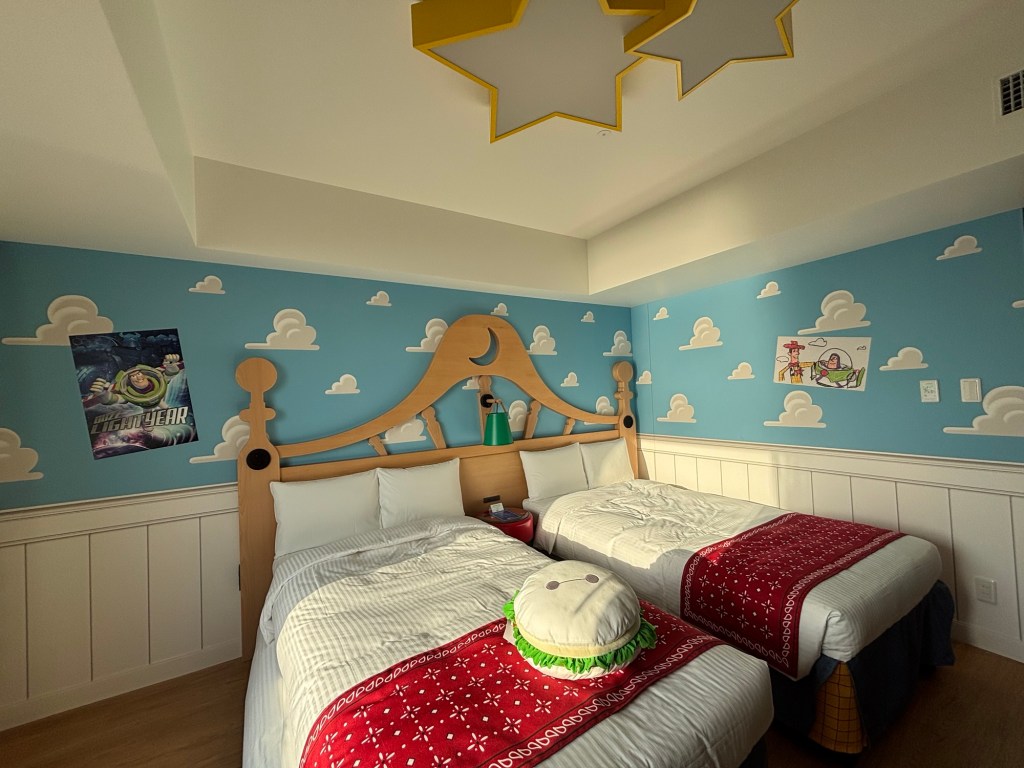 青い壁に雲の模様が描かれた部屋に、2つのベッドとカラフルなクッションが置いてある。壁には『バズ・ライトイヤー』のポスターとイラストが飾られている。
