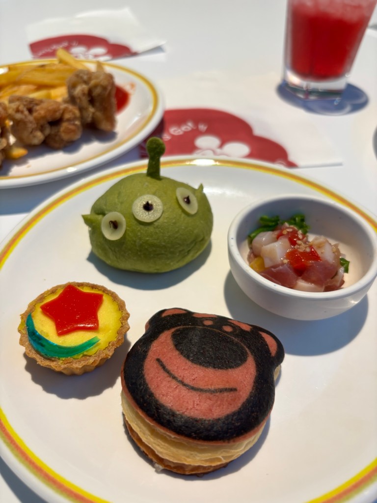 色とりどりのキャラクターを模したデザートの盛り合わせ。緑のエイリアン型のスイーツ、スターの飾りがのったカスタードタルト、クマの顔が描かれたクリームサンド、そしてサラダが盛り付けられた小皿が含まれている。