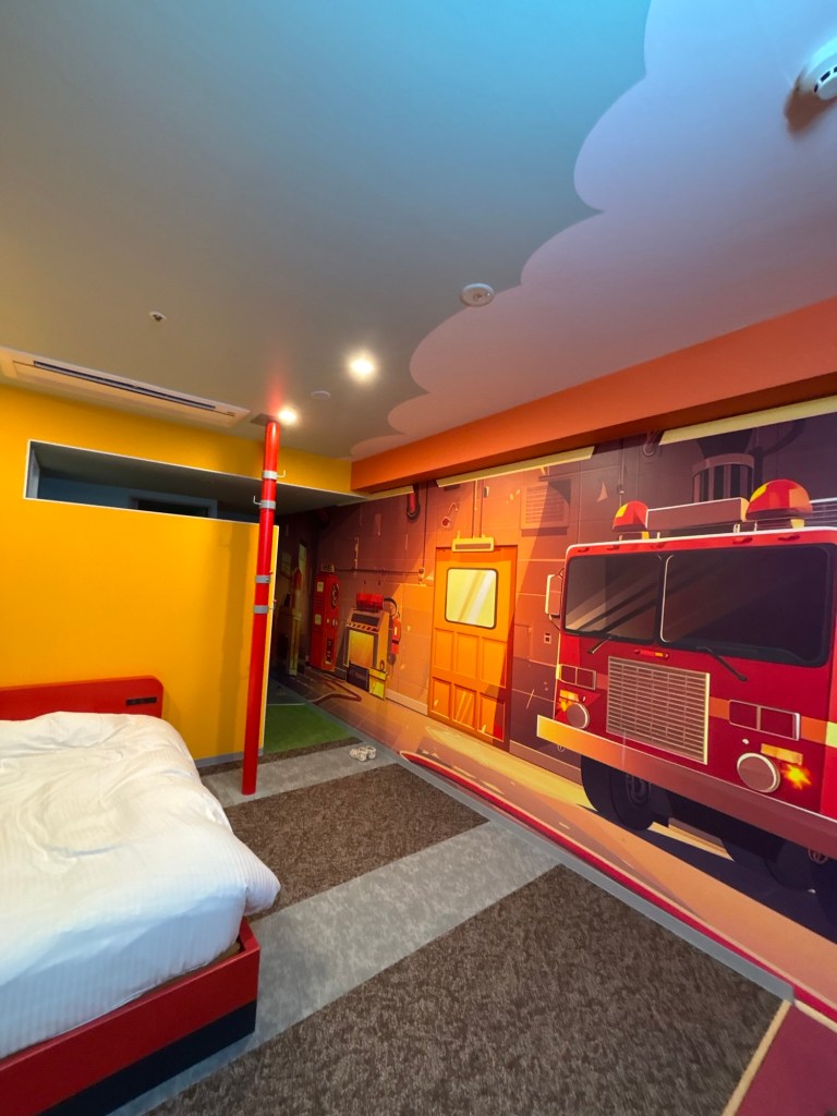 消防車の壁画が描かれたカラフルな子供部屋の内部。赤いベッドと整理されたスペースが特徴。