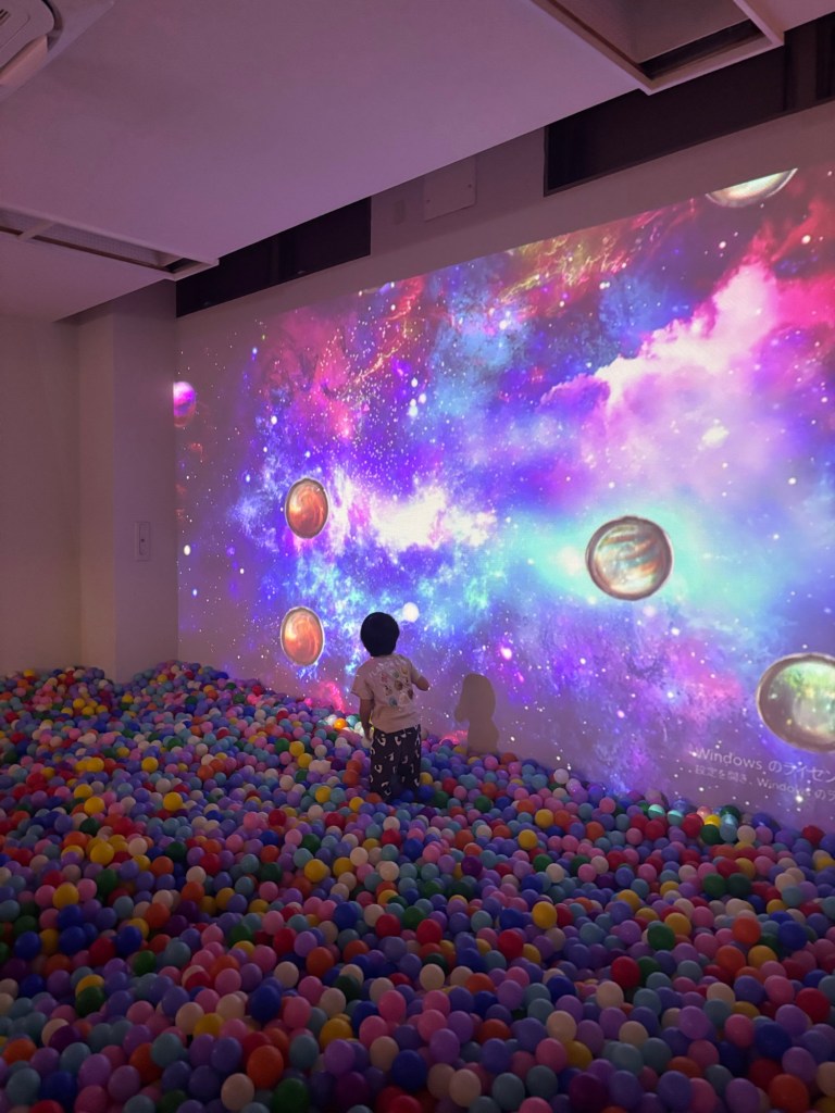 カラフルなボールプールの中に立つ子供と、宇宙の映像が投影された壁。