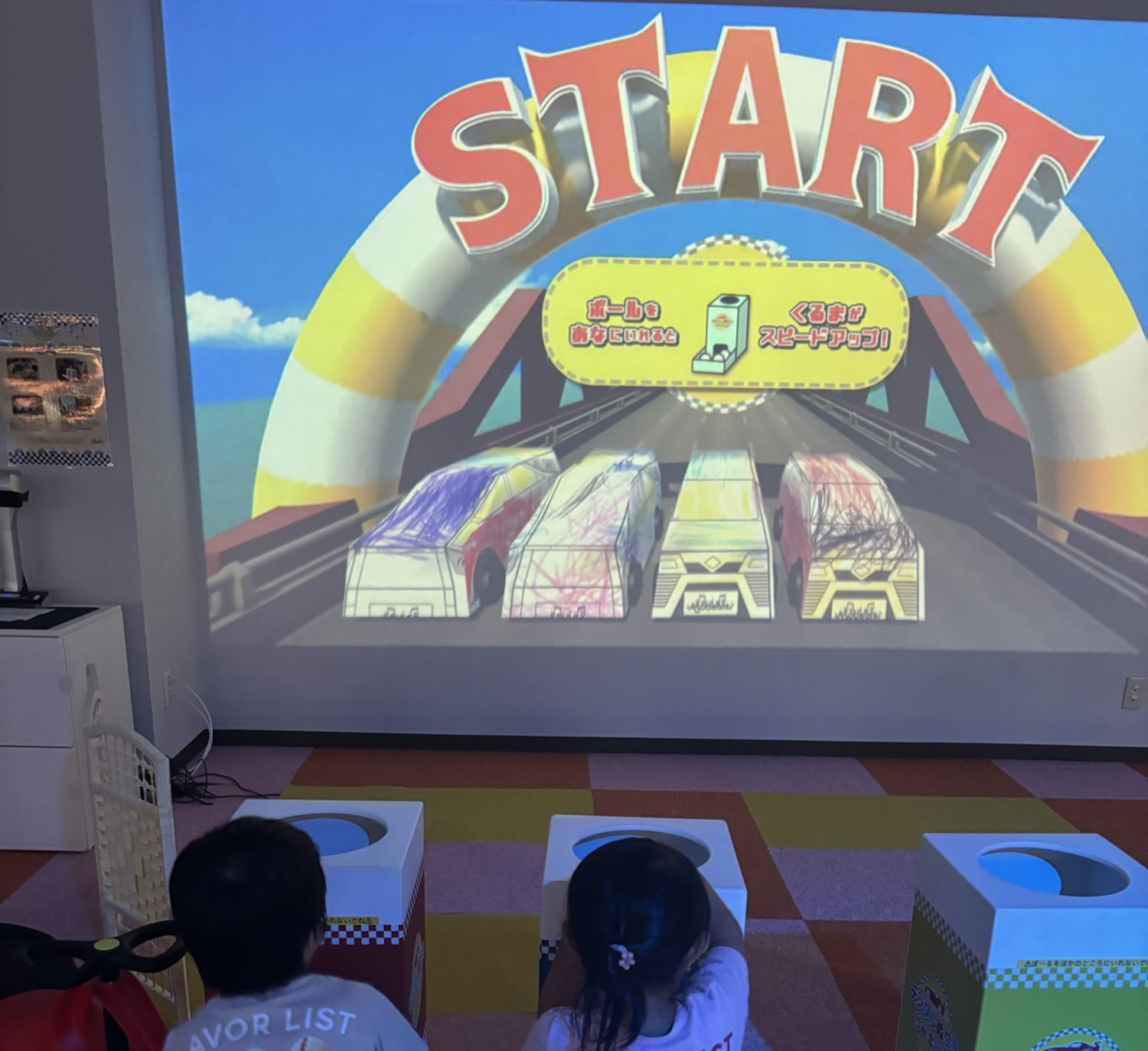 img_4373 子供たちがレースのゲームを楽しんでいる様子。画面にはカラフルな車が描かれたスタートの文字と一緒に表示されている。