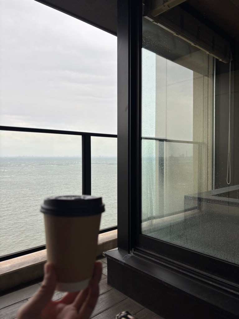 窓の外を見ながらコーヒーを持つ手。背景には海と曇り空が広がっている。